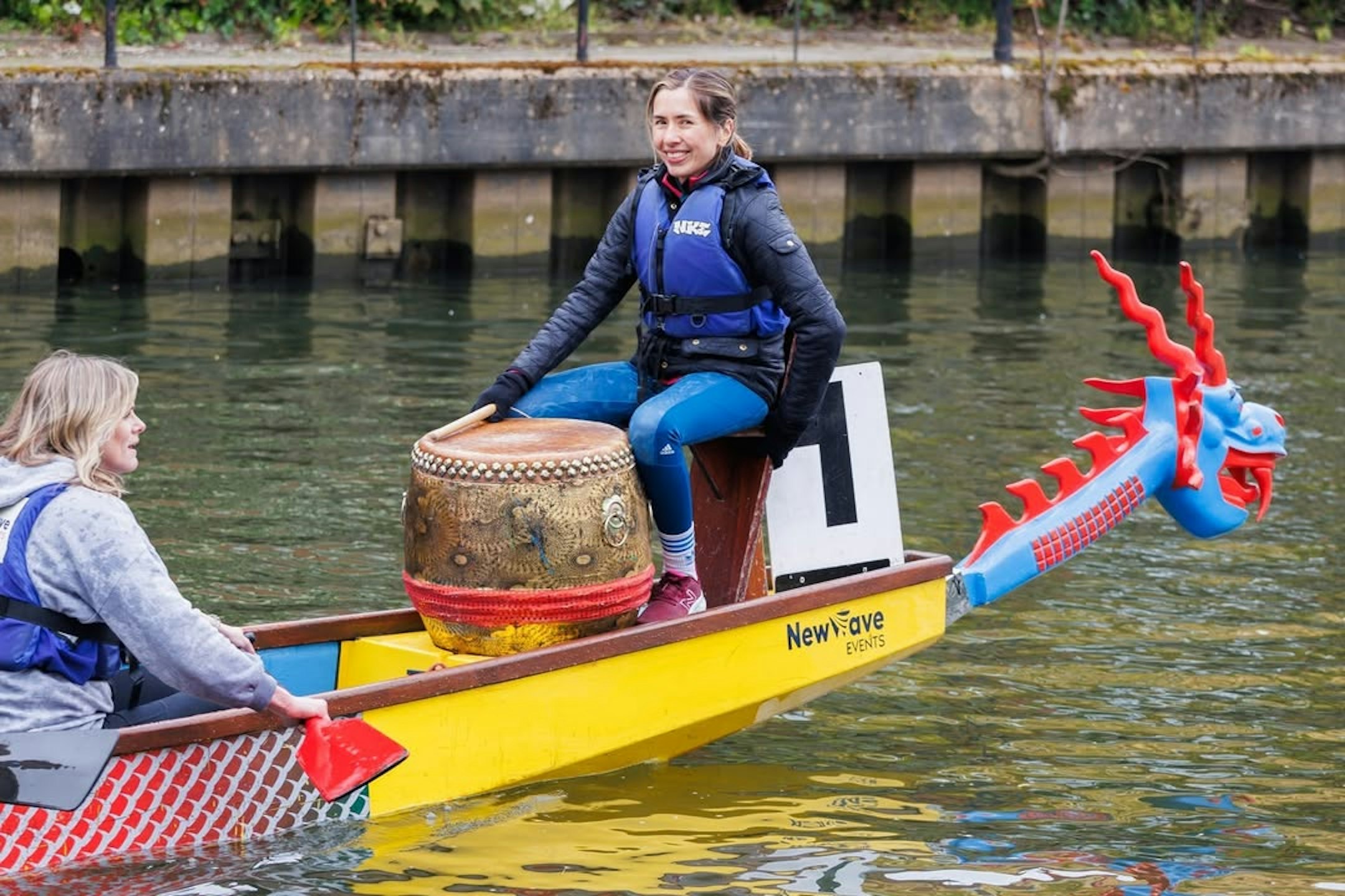 A female Candour colleague, wearing a buoyancy aid, is sitting at the front of a dragon boat on a river. There is a large drum in front of her.