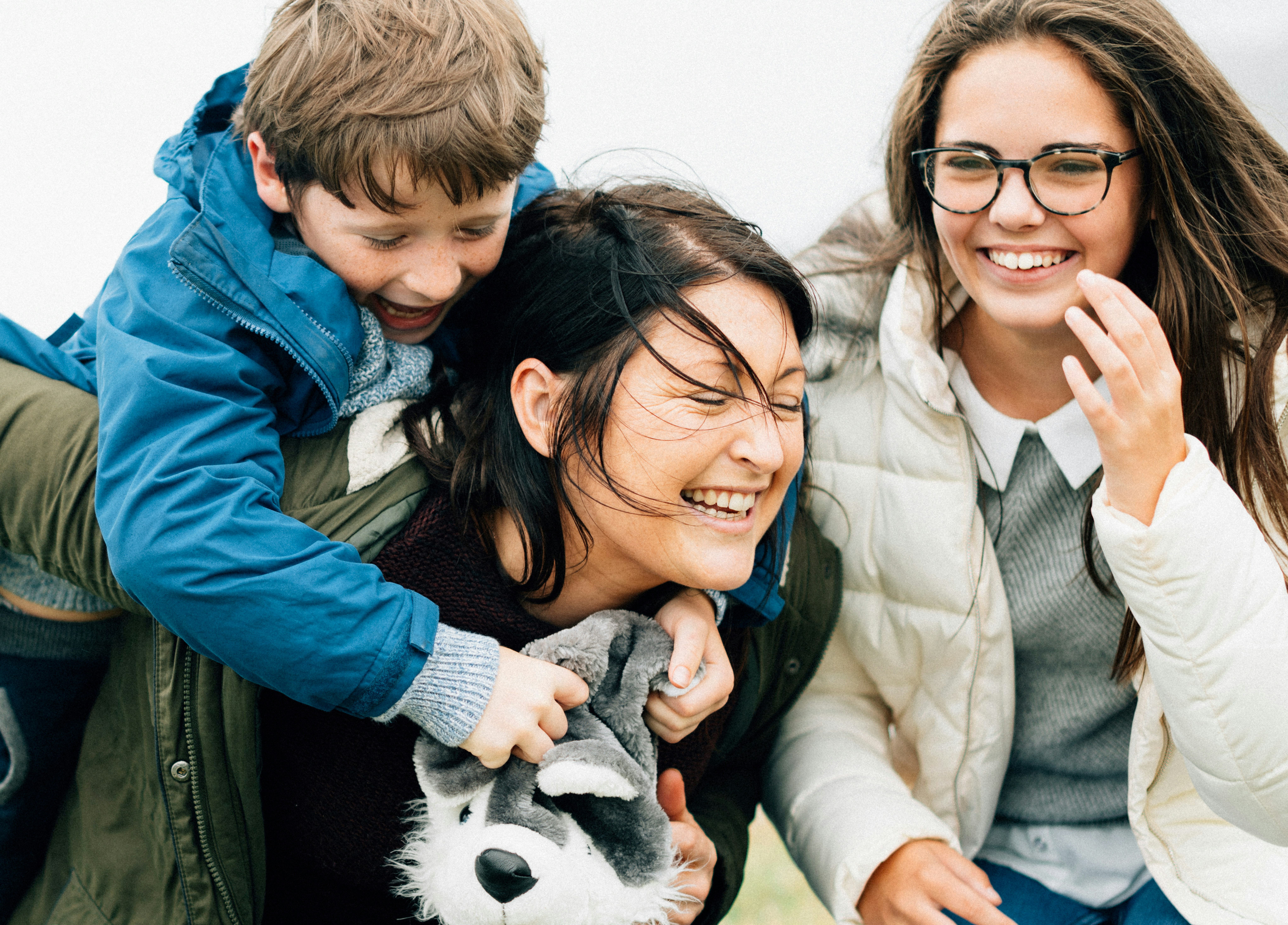 A foster carer with two children. All three are smiling and having fun. The carer is carrying a boy aged about 8 on her back and is standing next to a teenaged girl who is wearing glasses