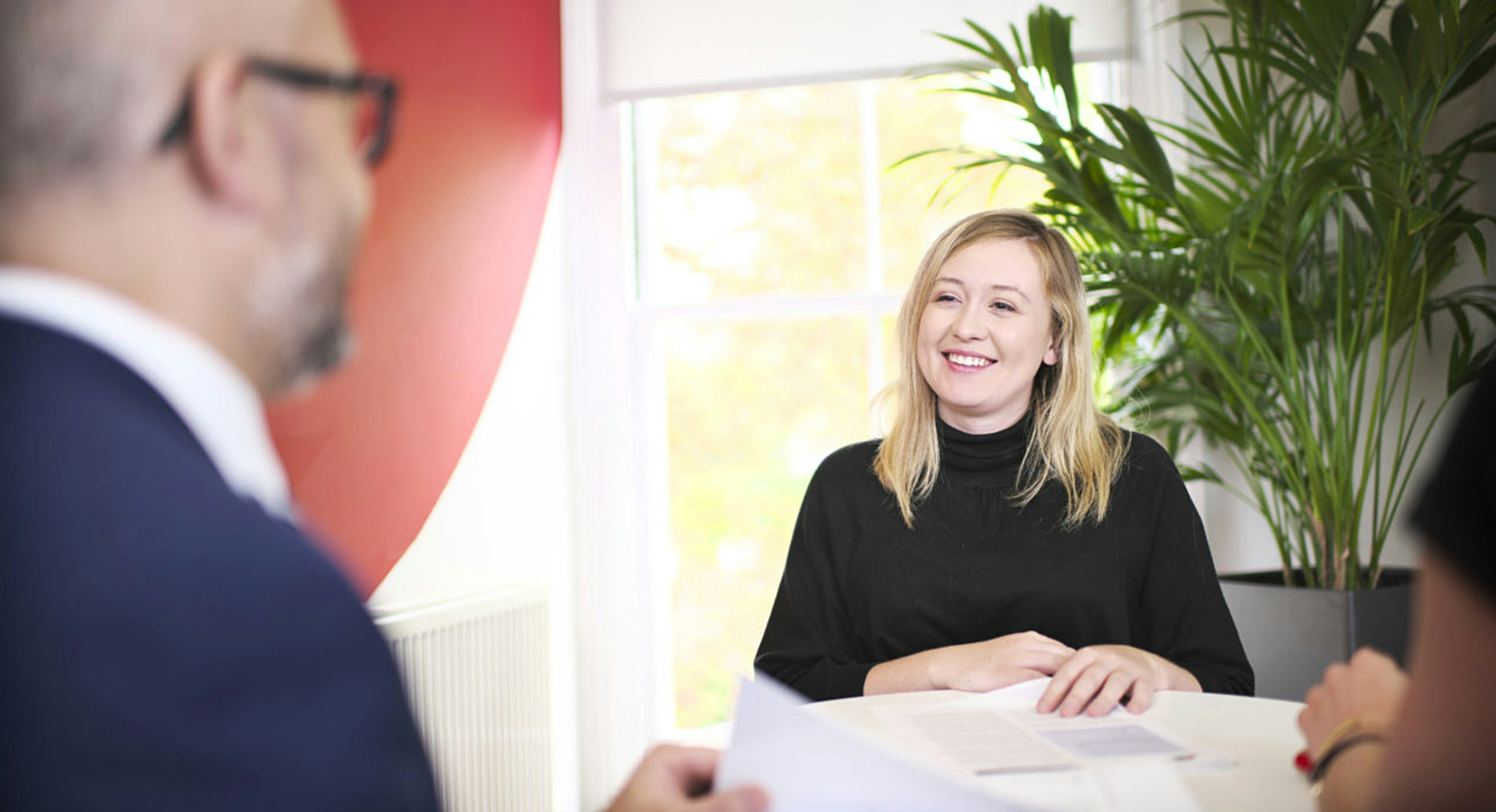 A smiling woman in a black turtleneck sits at a table during a professional meeting, engaging in conversation with two blurred figures in the foreground.