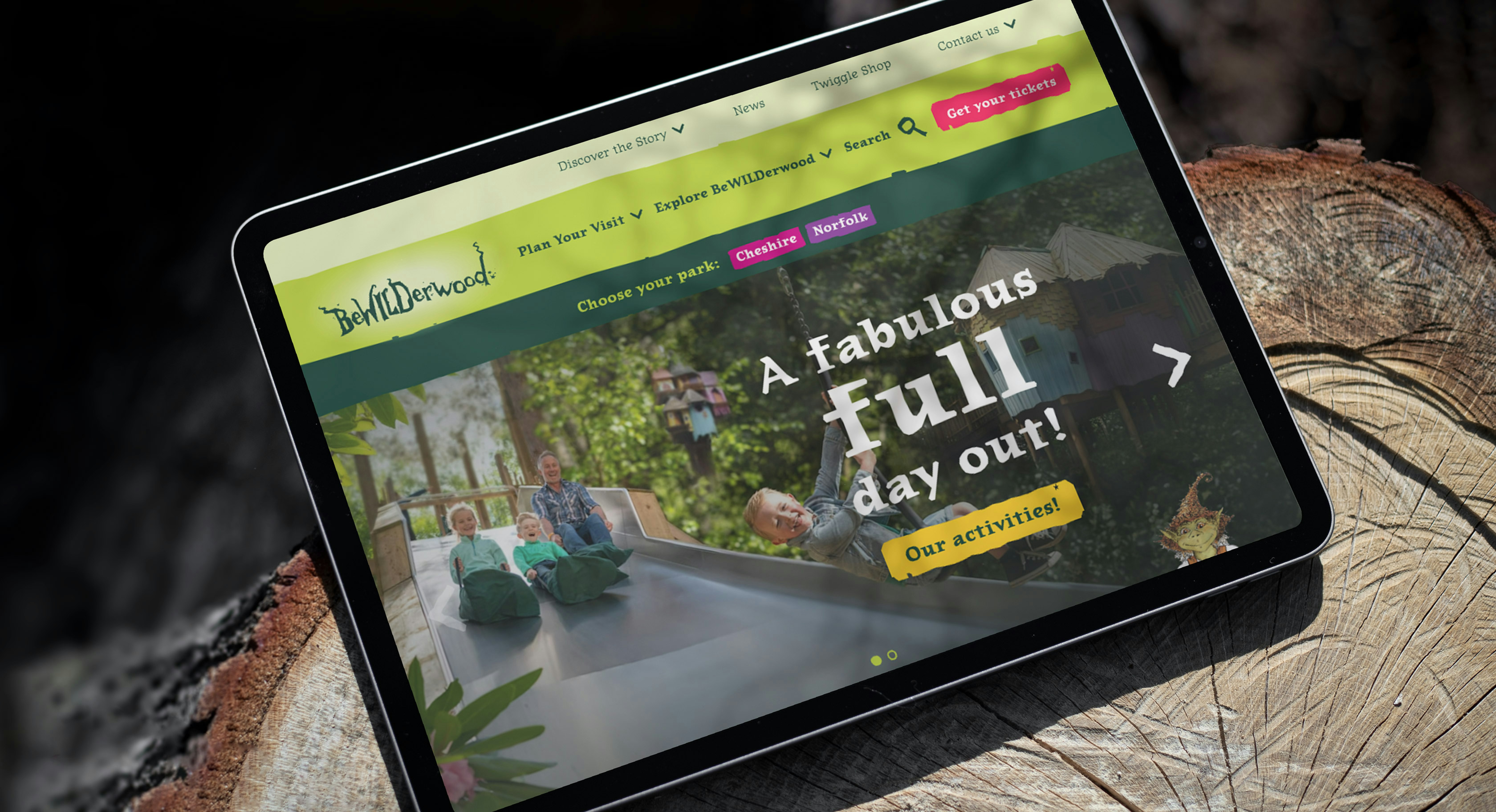 An ipad sitting on a log displaying the homepage of the BeWILDerwood website
