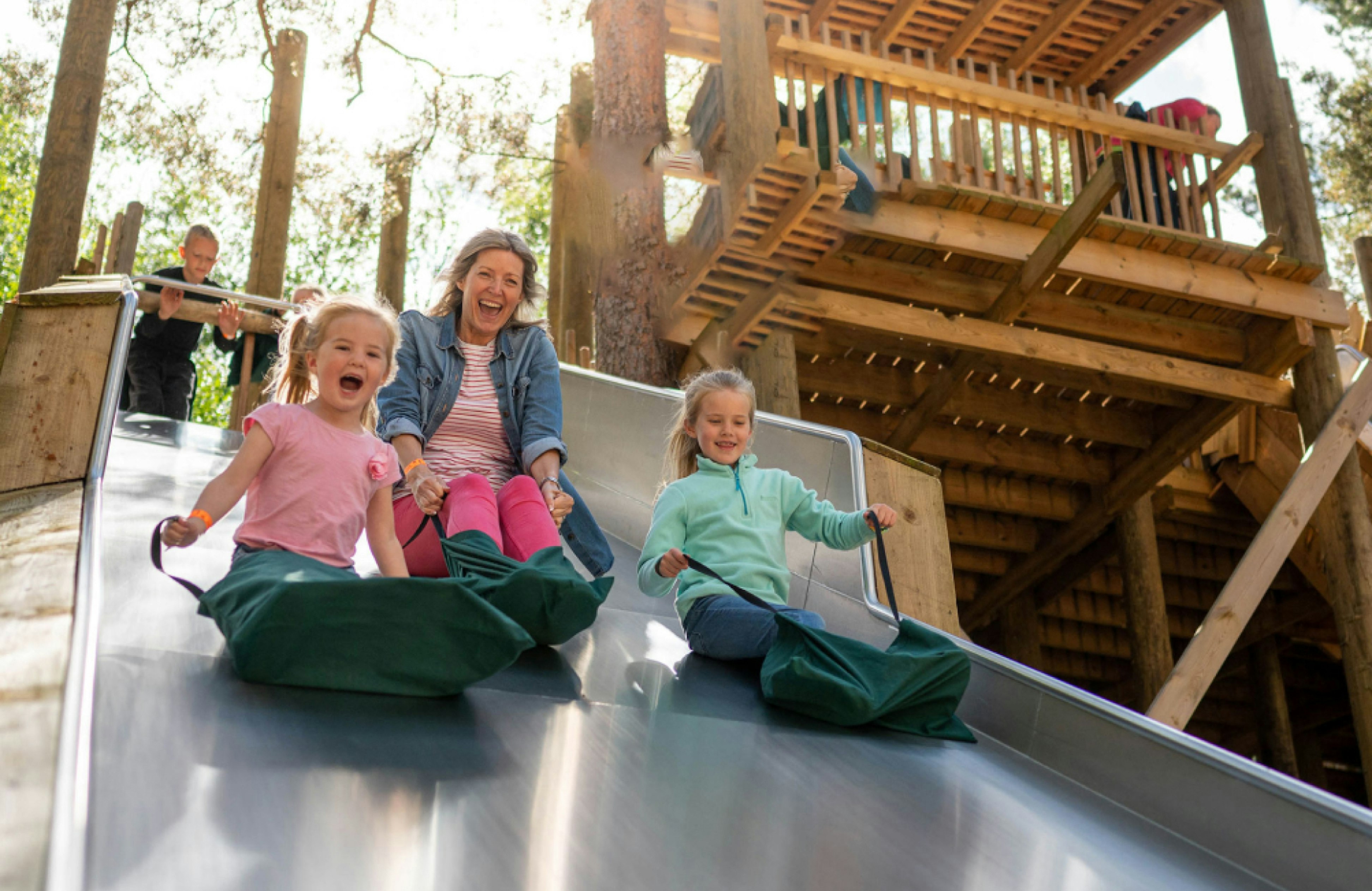 A mother and two young girls laughing as they slide down a metal slide on green mats, with a wooden treehouse structure in the background.