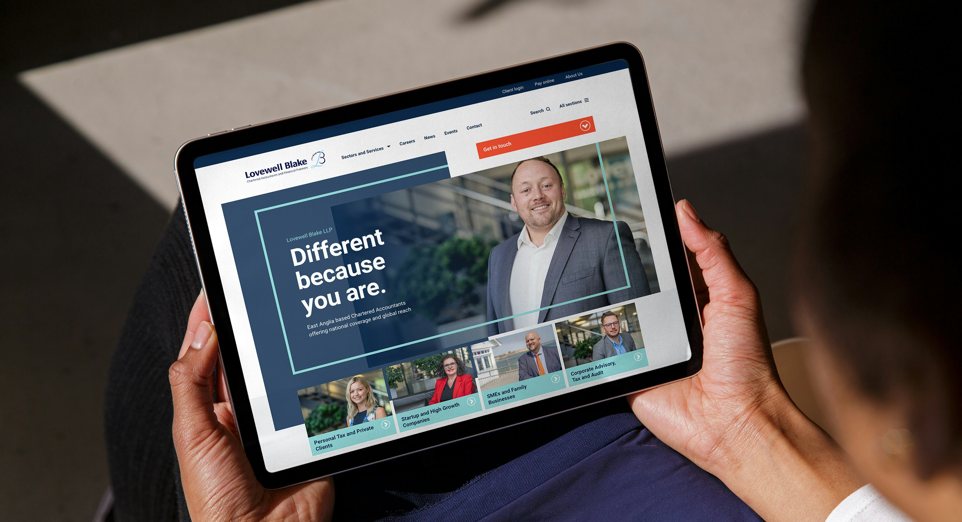 Two hands holding an iPad tablet computer displaying the homepage of the Lovewell Blake website
