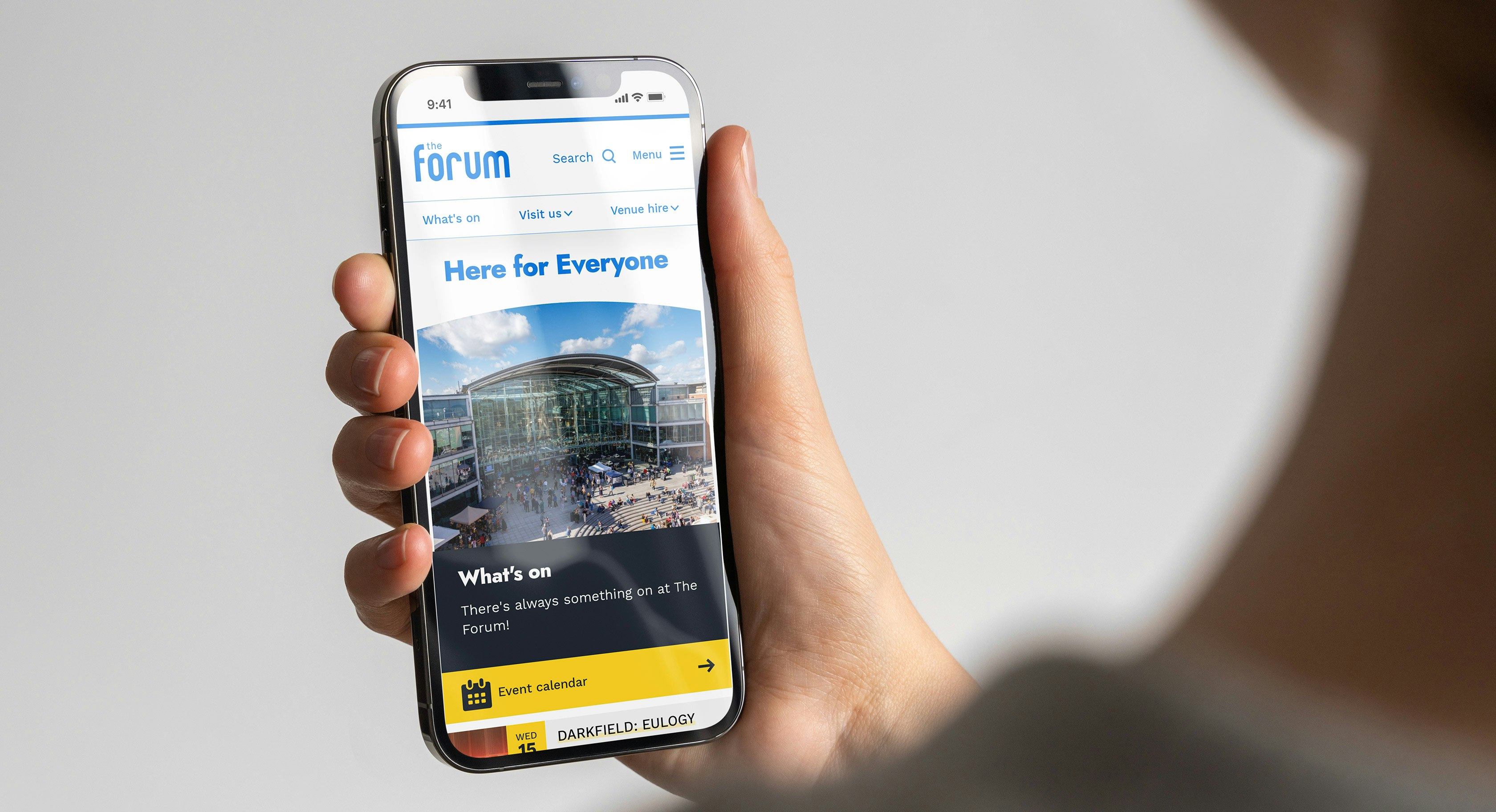 A hand holding a mobile phone that is displaying the homepage hero section of the The Forum website