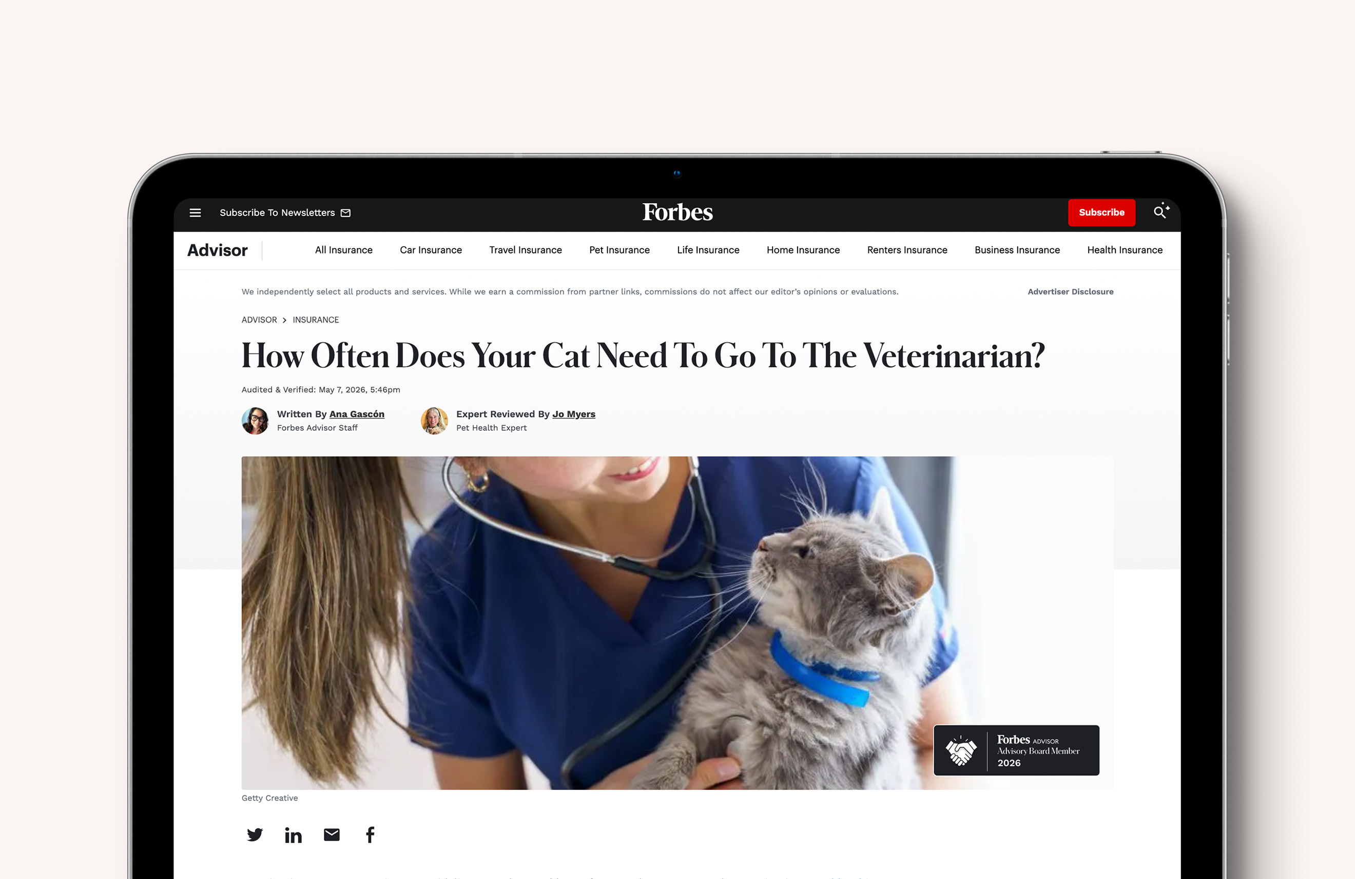 A tablet displays a Forbes Advisor article titled “How Often Does Your Cat Need To Go To The Veterinarian?” with a photo of a veterinarian examining a gray cat wearing a blue collar.