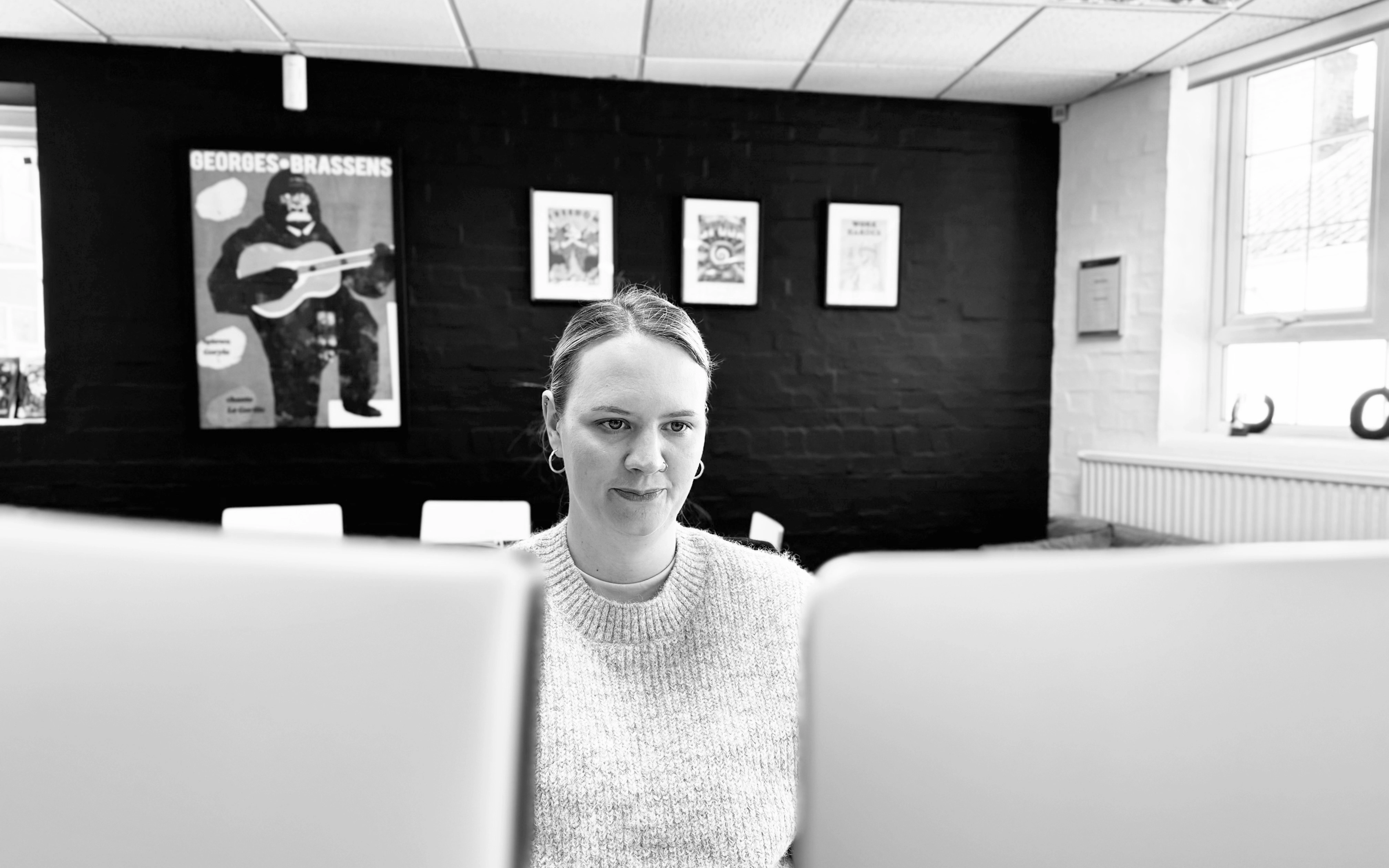 Black and white photo of a person sitting at a desk, partially hidden behind two computer screens, in a modern office with framed posters on a dark brick wall in the background.