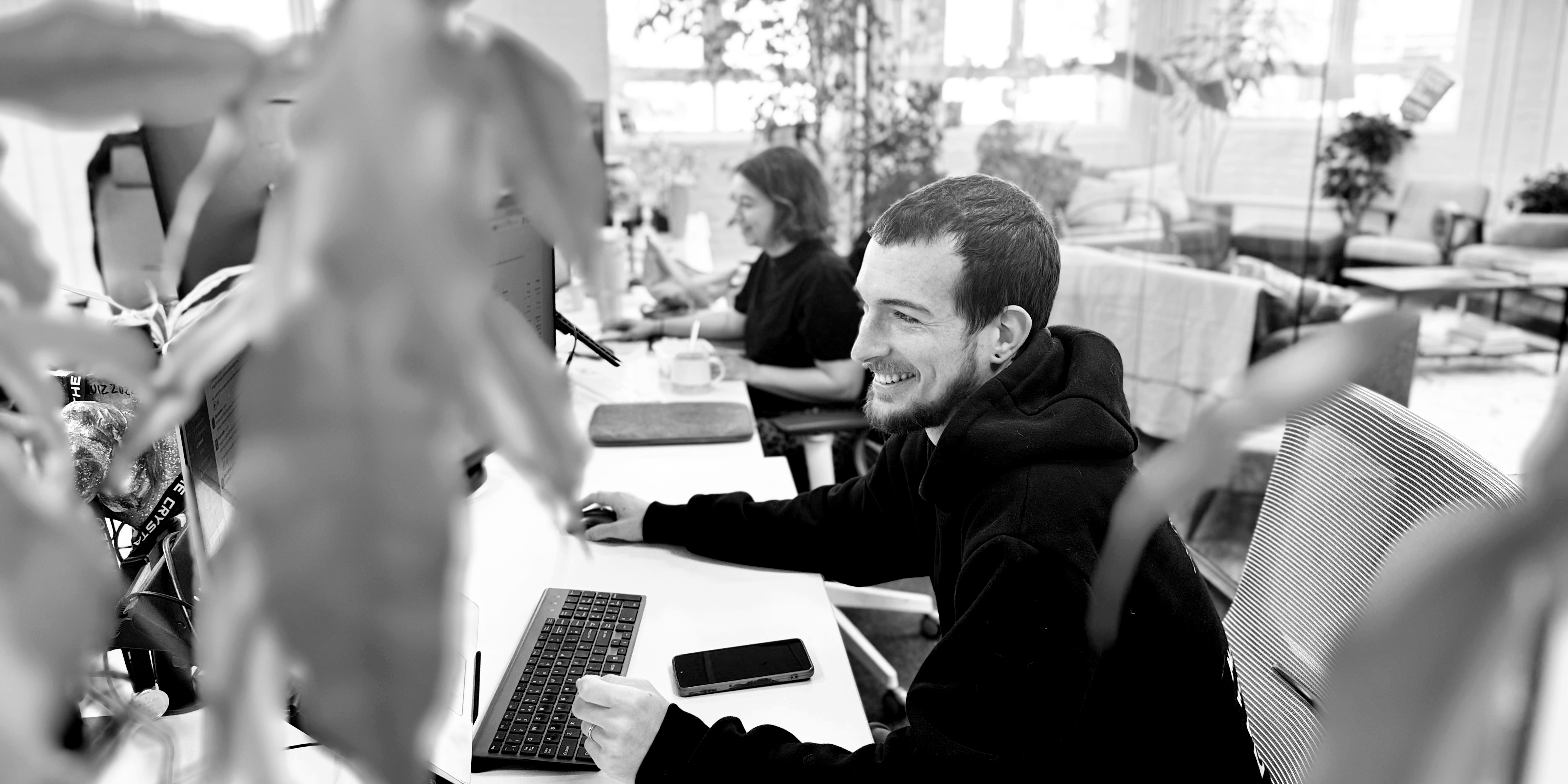 Black and white photo of a modern open‑plan office with two people working at computers, their faces blurred, seen through out‑of‑focus plant leaves in the foreground.