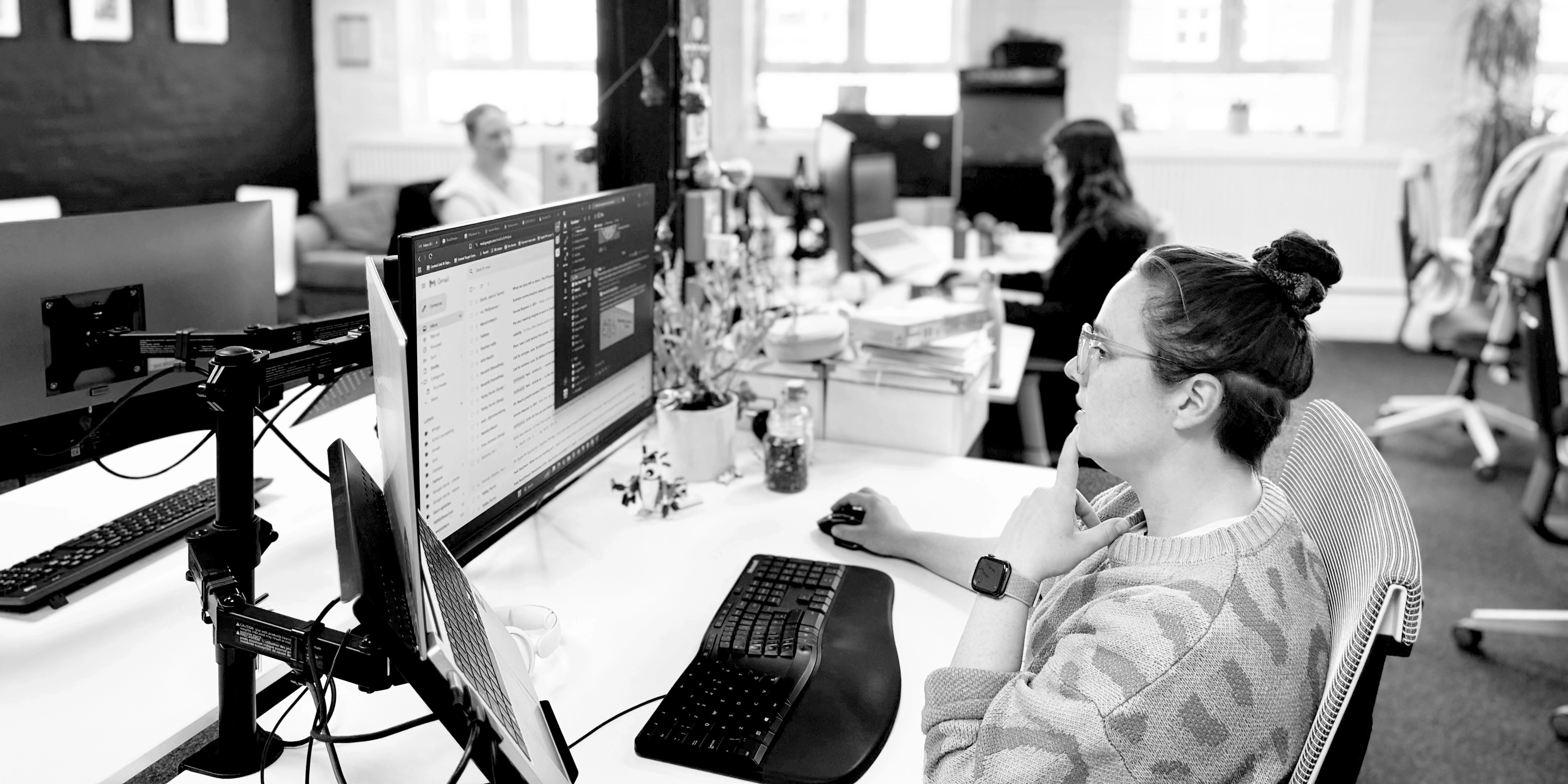 A black-and-white image of a modern, open-plan office space. In the foreground, a woman wearing glasses, a patterned sweater, and a smartwatch sits at her desk, looking thoughtfully at a large computer monitor with one hand resting on her chin. Her desk features an ergonomic keyboard, mouse, a laptop mounted beside the monitor, and small potted plants. In the background, other colleagues are seated at their desks working on laptops.
