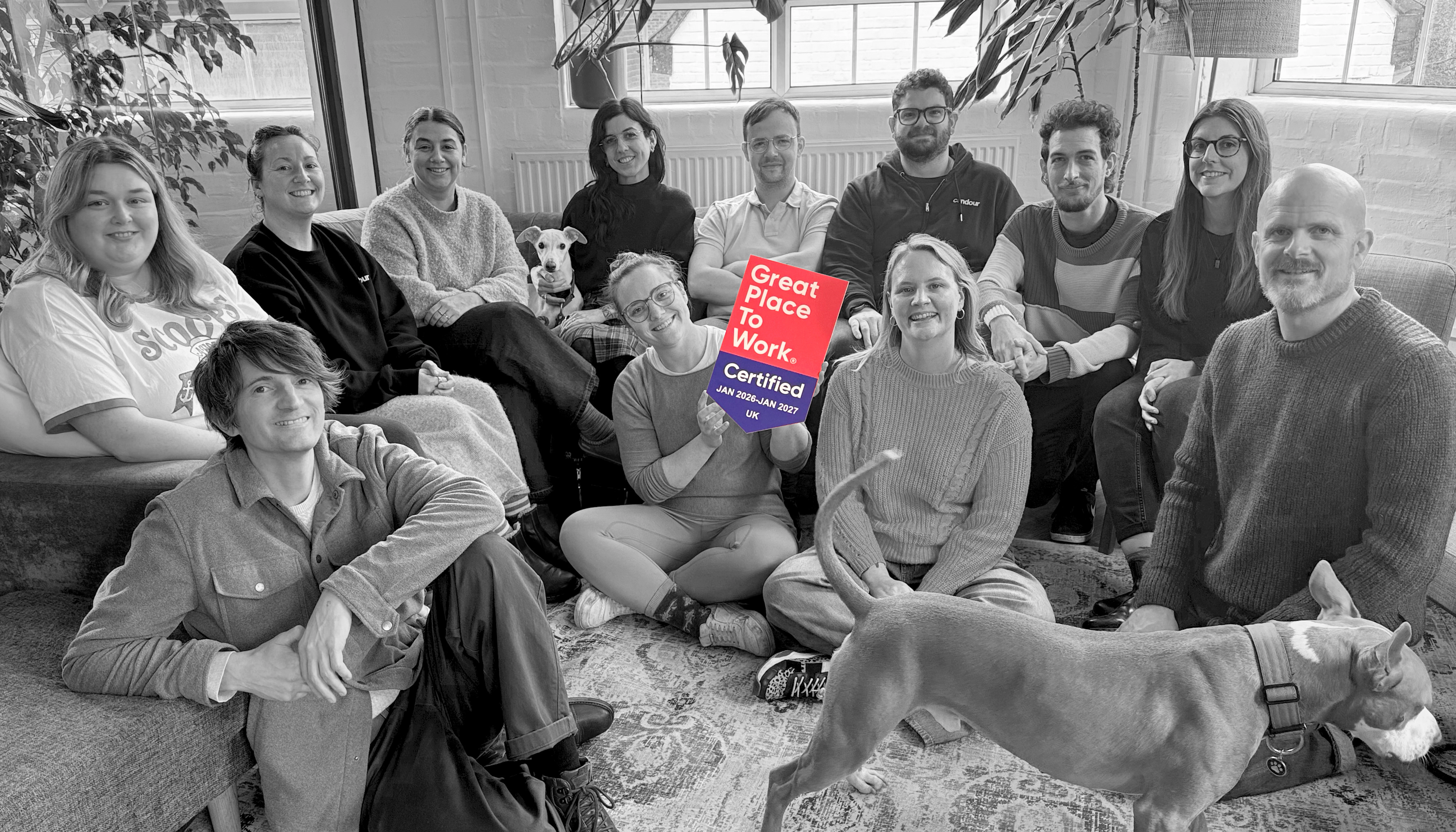 A black and white group photo of the Candour team sitting together in a casual office setting. In the center, a team member holds a brightly colored red and blue plaque that reads