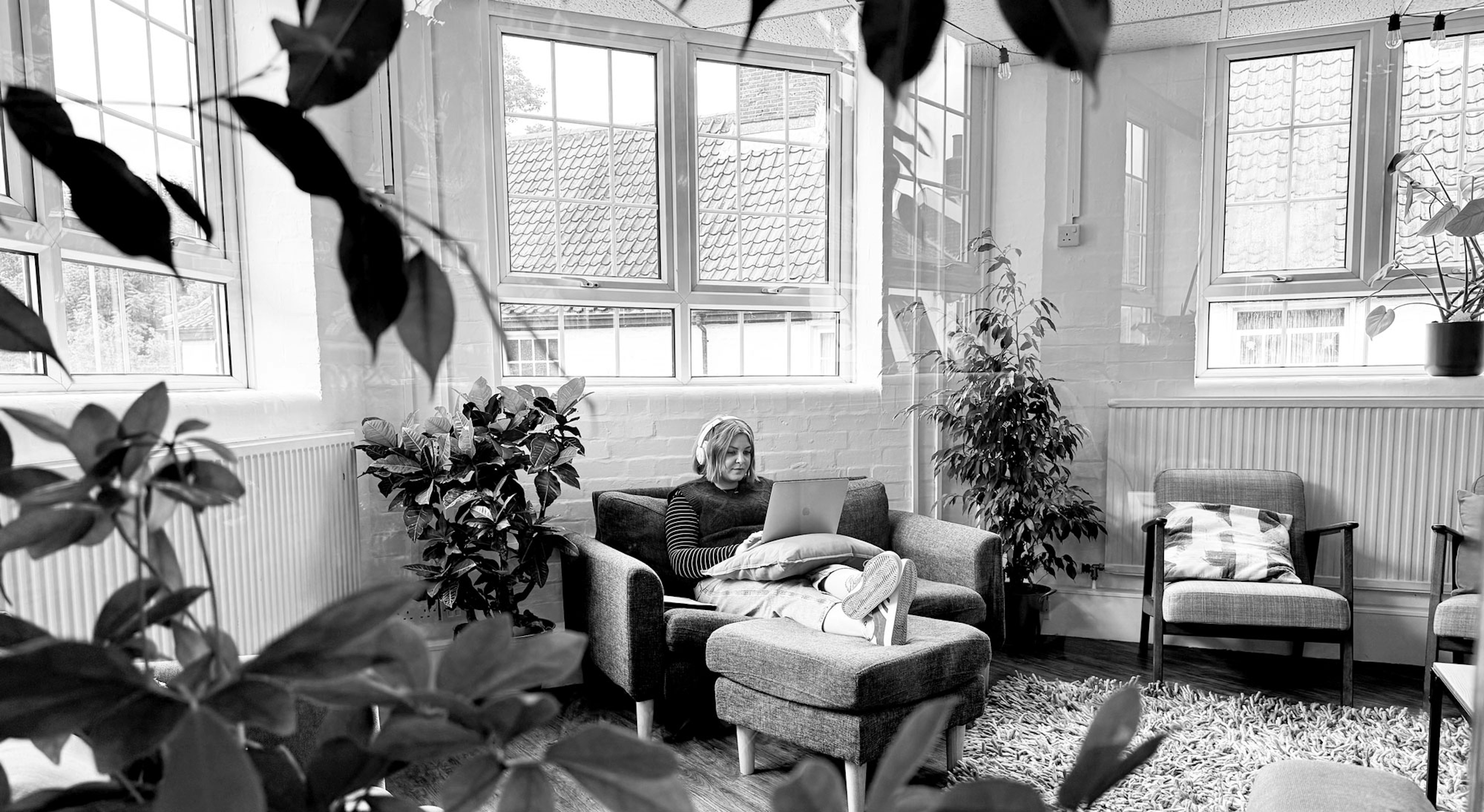 Jessica Bettis sitting on a sofa while working on her laptop. Her feet are resting on a pouffe and she is wearing headphones.