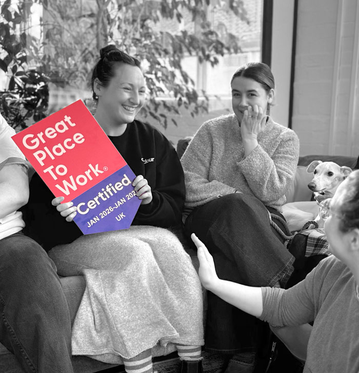 A black-and-white photo of three smiling women sitting together on a sofa in a casual office setting, with a whippet dog peering out from behind the woman in the centre. The woman on the left holds up a brightly coloured, shield-shaped sign that stands out against the monochrome background. The red and blue sign reads: 