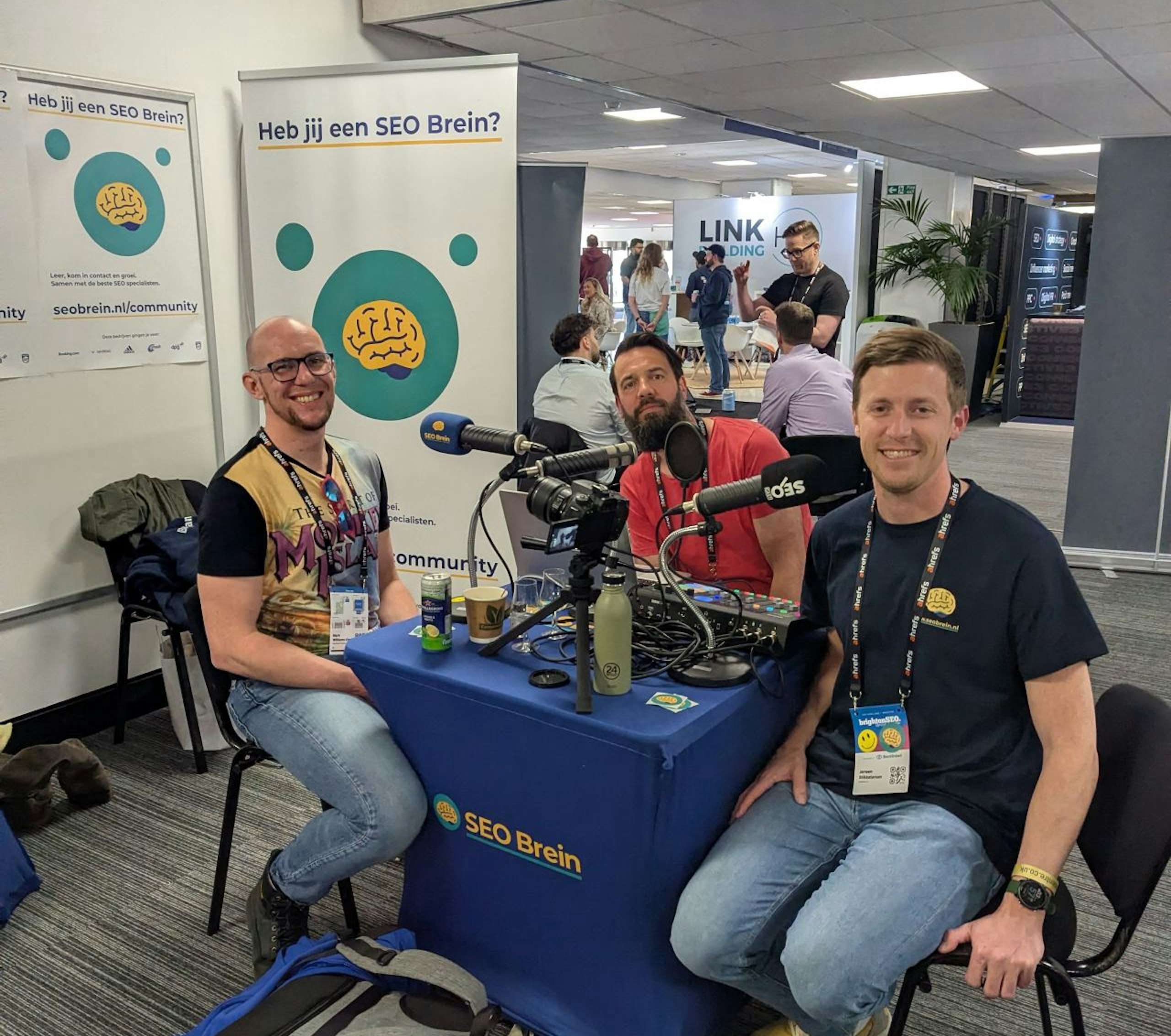 Mark Williams-Cook (still in his Monkey Island t-shirt) sitting down with two other smiling gents in front of microphones while being interviewed for a podcast at BrightonSEO