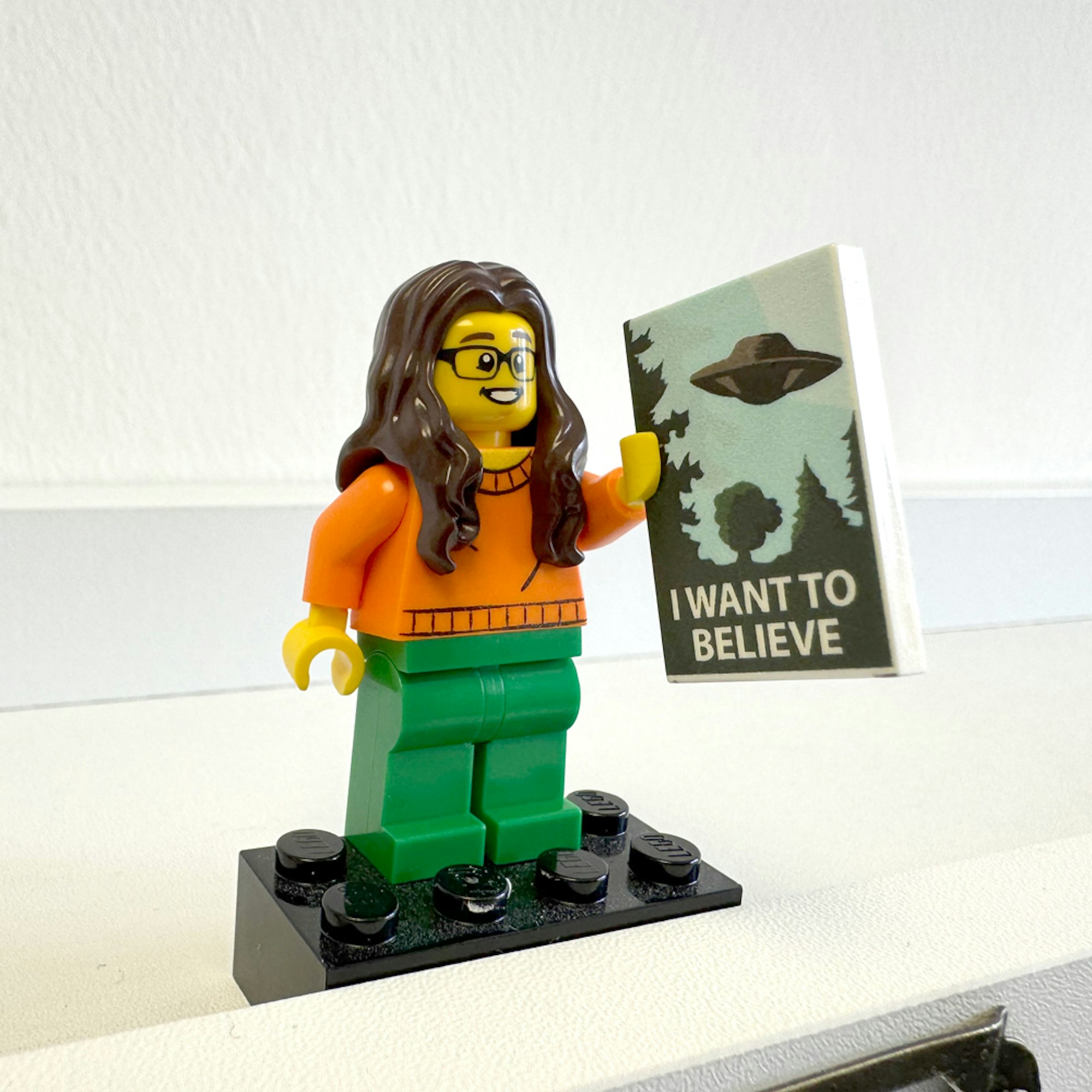 A closeup of Sammy Marshall's lego figure. She is holding a book with an image of a UFO, entitled 'I want to believe' and wearing green trousers and an orange jumper.