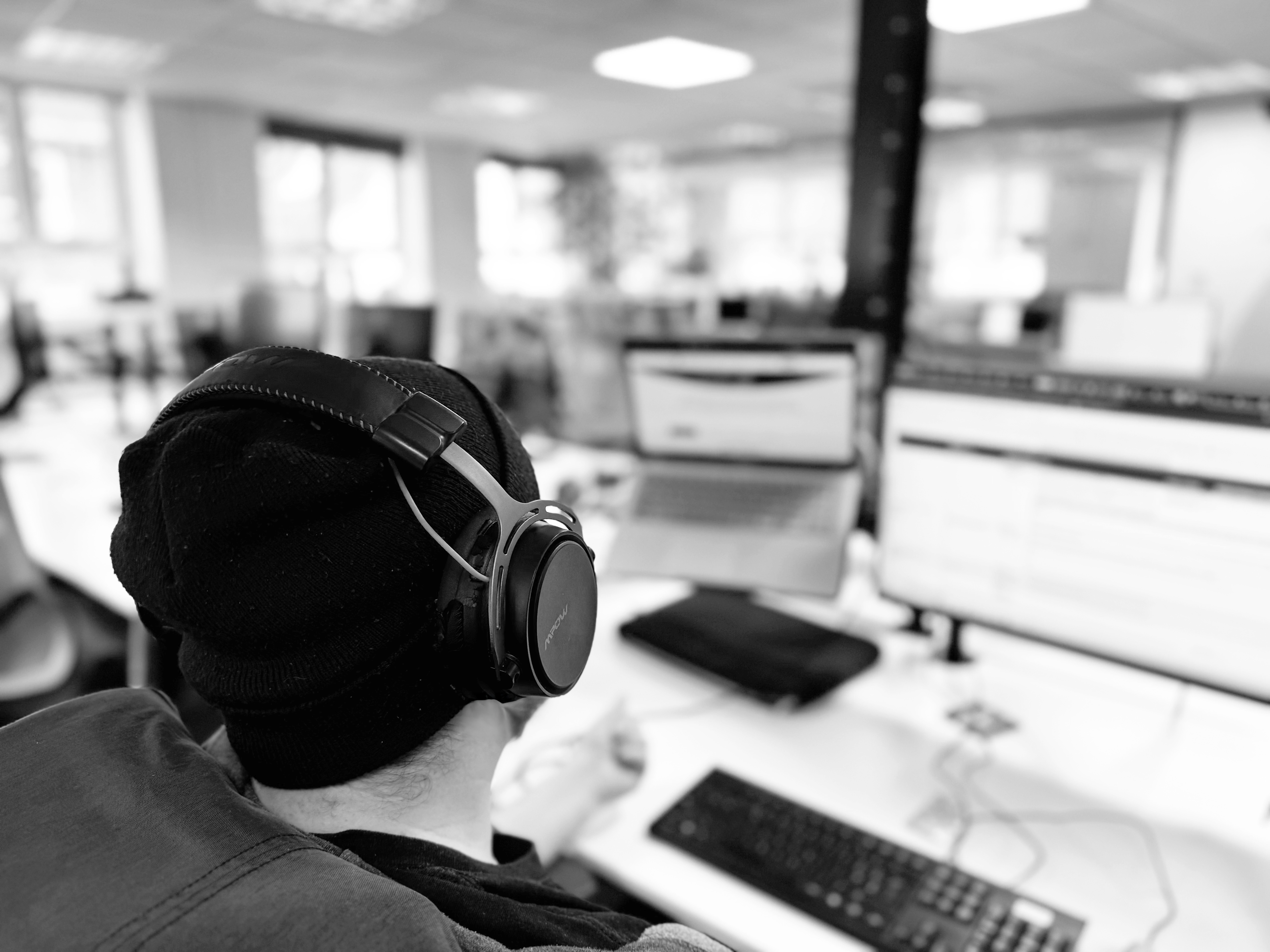 the back of a man's head as he works at his desk in front of computer screens. He is wearing  a beanie hat and headphones.