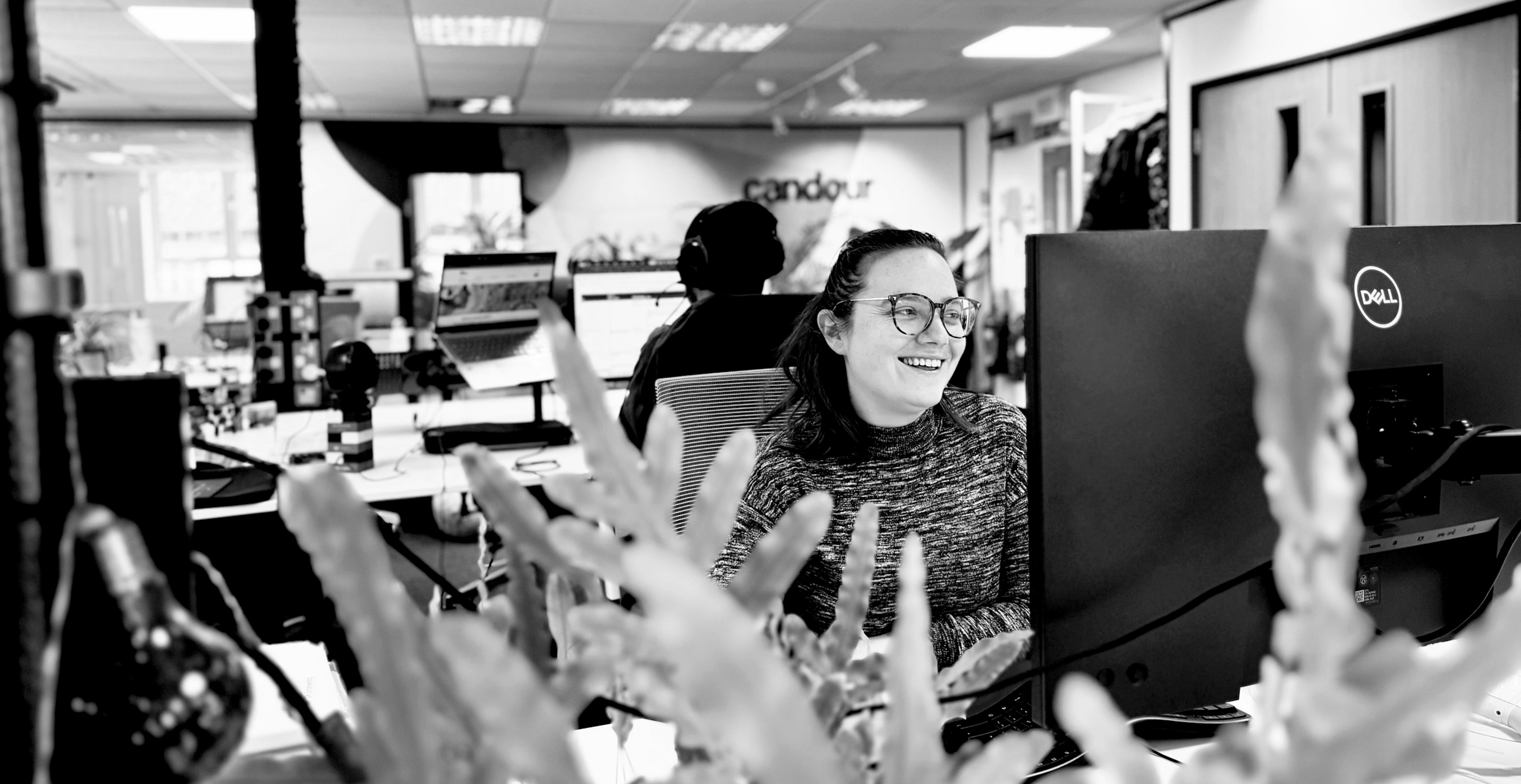 A young woman is sitting at her desk working in an office, she is smiling. There are the leaves of a plant in the foregrounds and a co-worker sat behind facing the other way