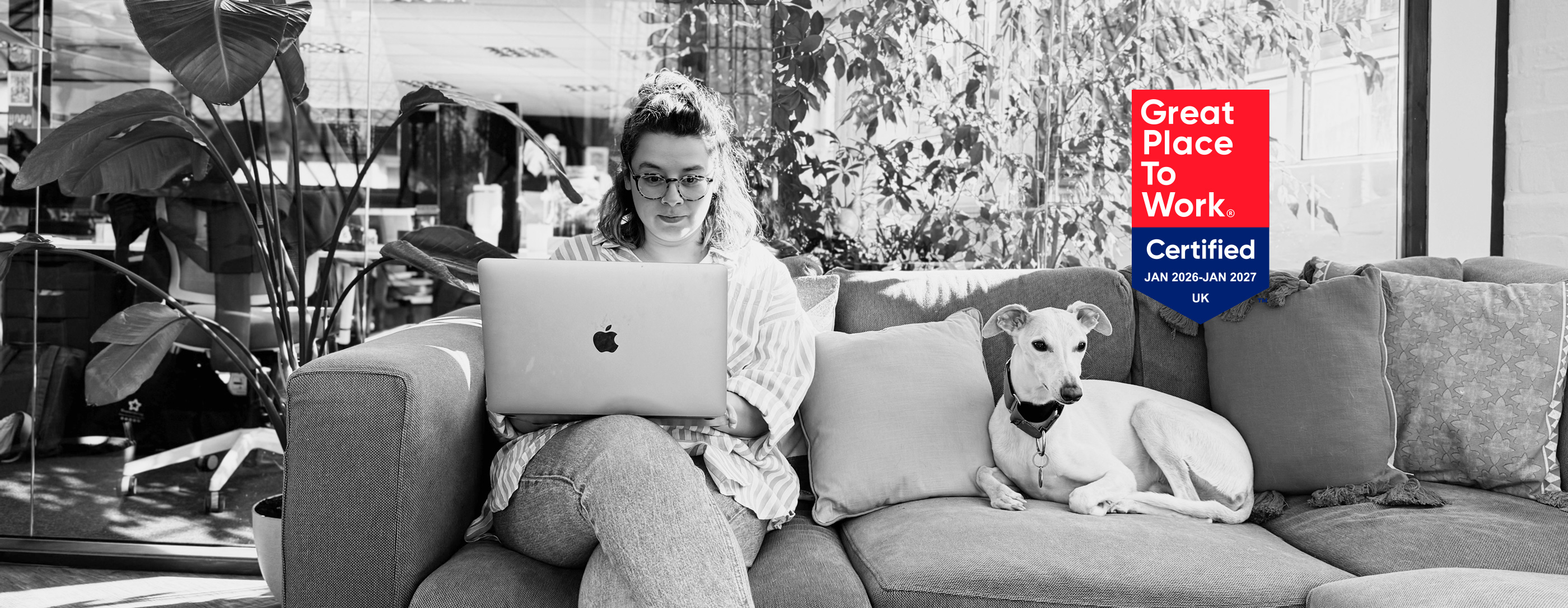 A black and white photograph of a woman wearing glasses sitting on a couch and working on an Apple laptop, accompanied by a light-colored dog resting next to her. A vibrant red and blue 