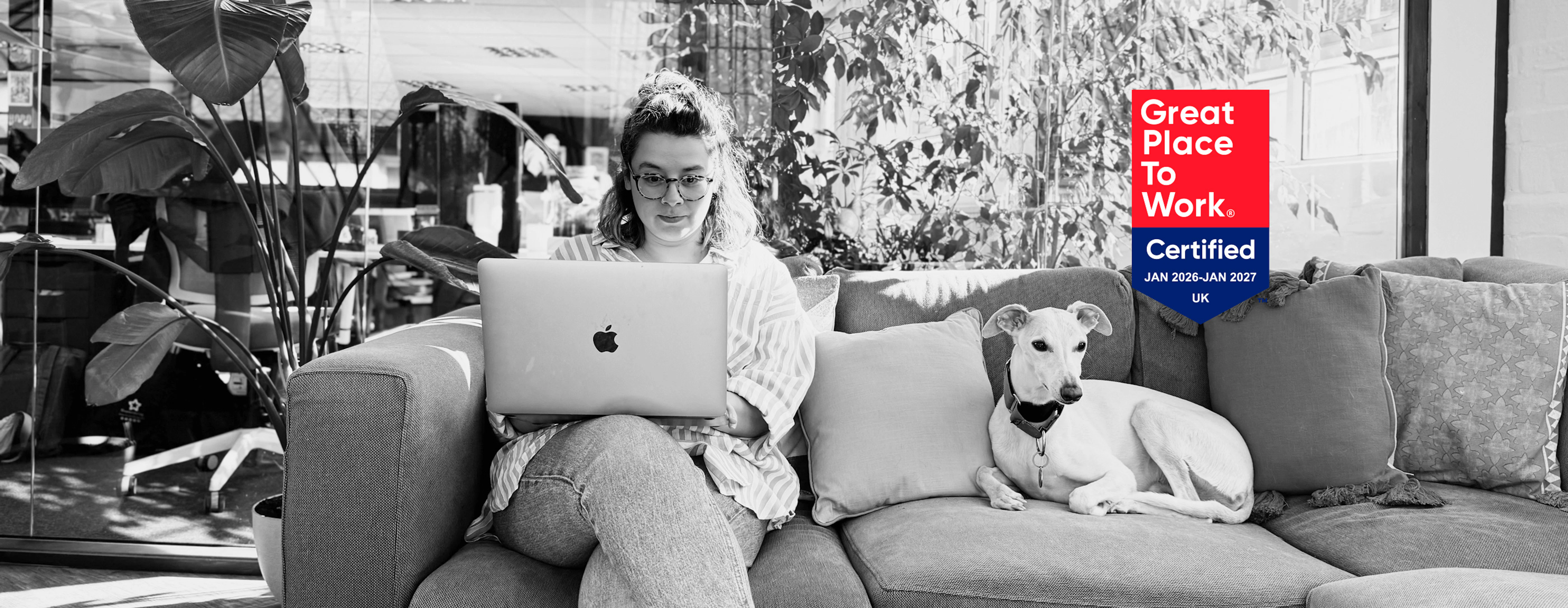 A black and white photograph of a woman wearing glasses sitting on a couch and working on an Apple laptop, accompanied by a light-colored dog resting next to her. A vibrant red and blue