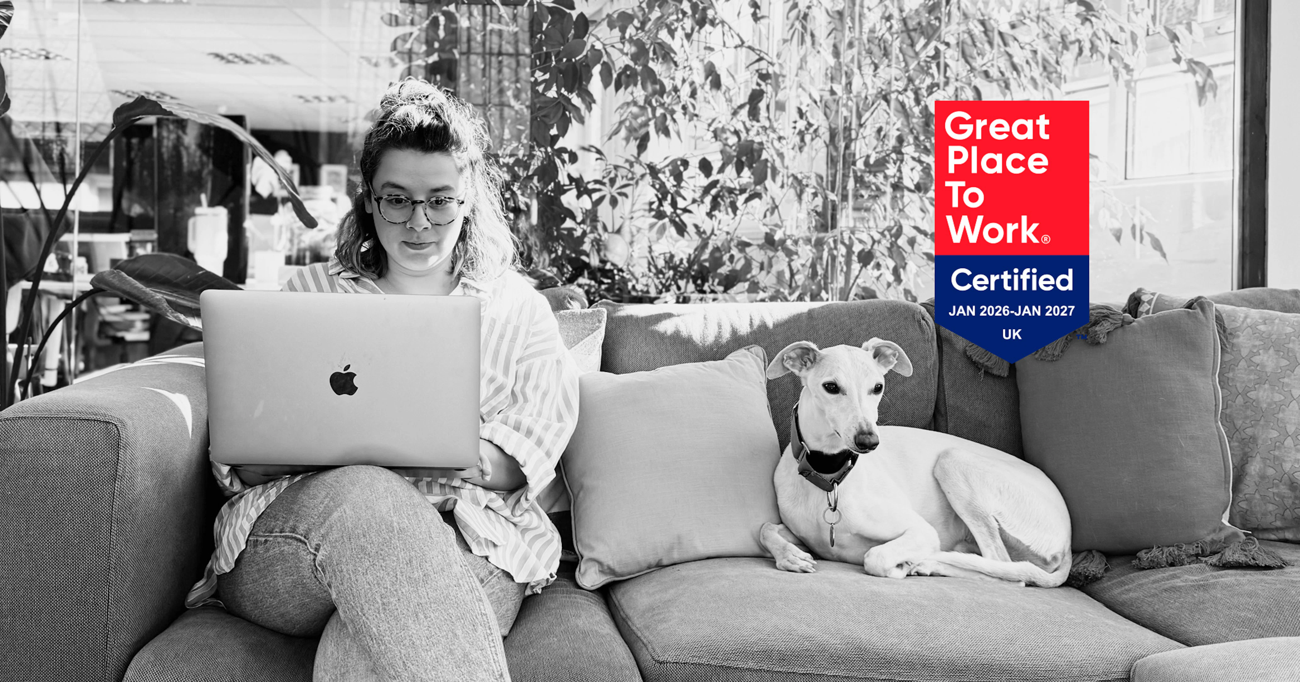 A black and white photograph of a woman wearing glasses sitting on a couch and working on an Apple laptop, accompanied by a light-colored dog resting next to her. A vibrant red and blue