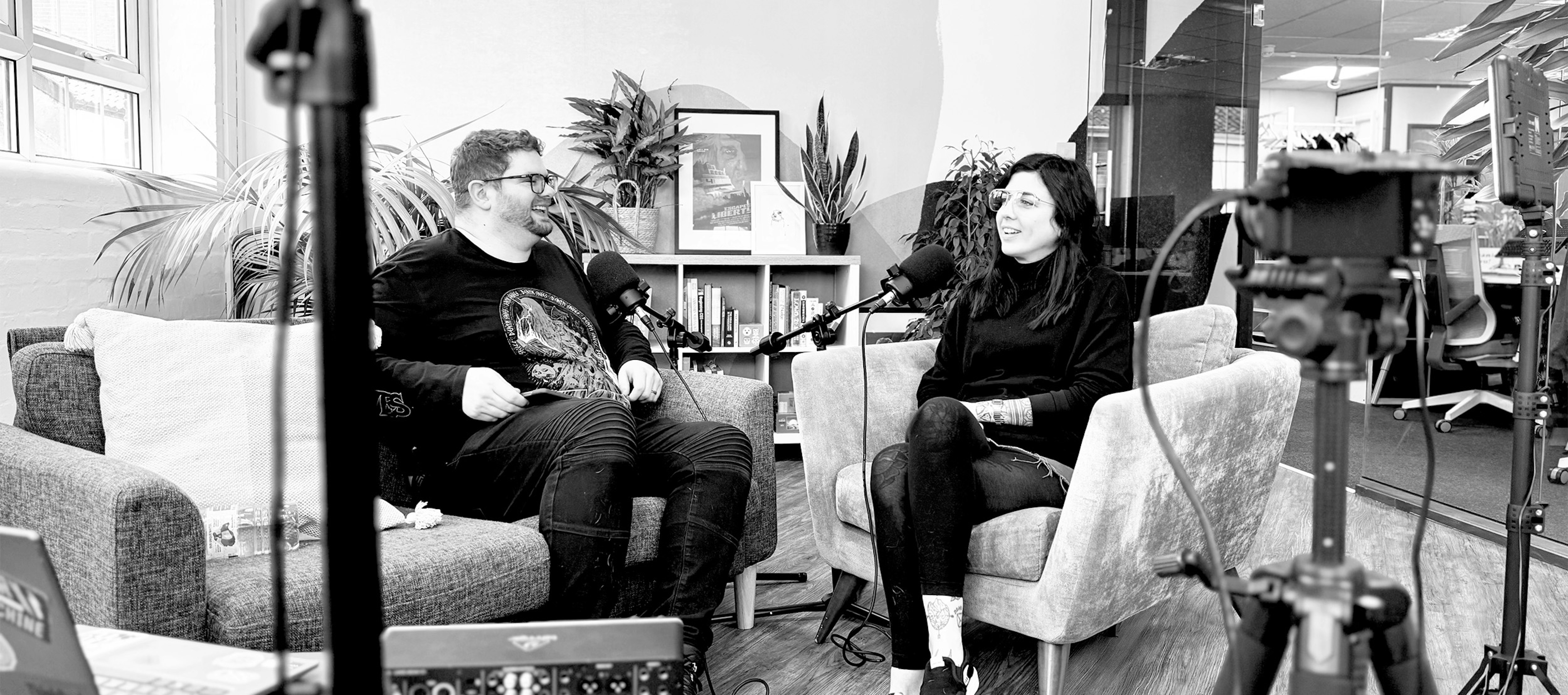 Jack Chambers-Ward and Tabby Farrar in conversation on two sofa chairs in an office break out room. They are speaking into microphones and a camera and lighting equipment can be seen in the foreground