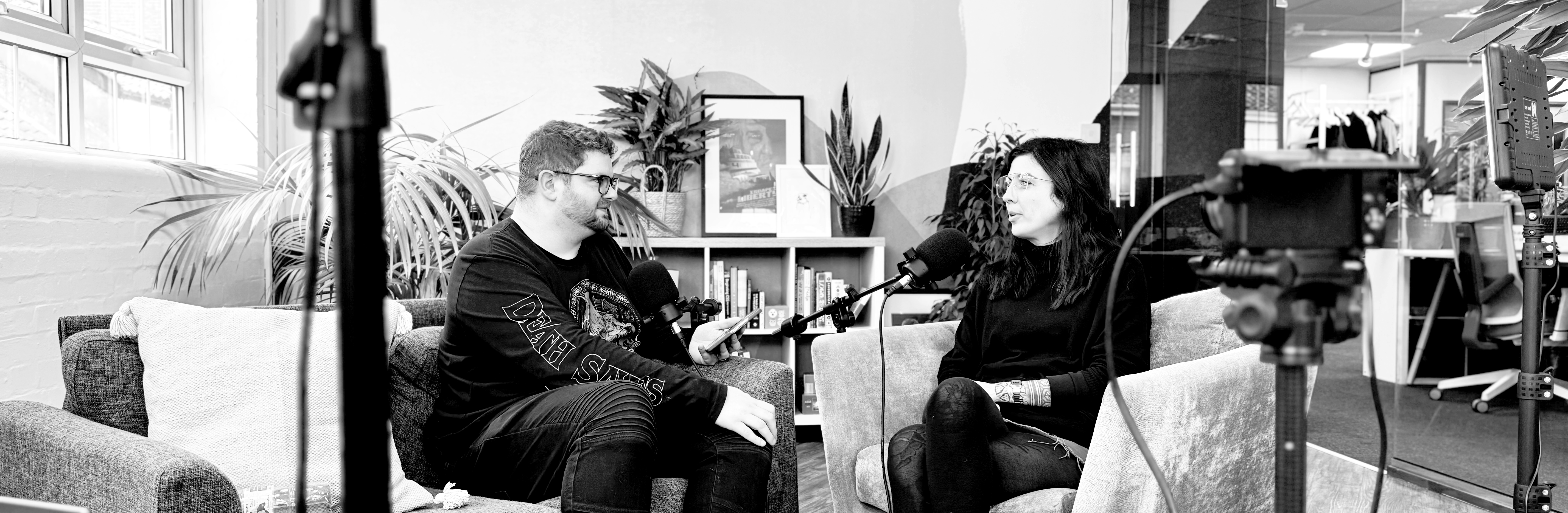 Jack Chambers-Ward and Tabby Farrar in conversation on two sofa chairs in an office break out room. They are speaking into microphones and a camera and lighting equipment can be seen in the foreground
