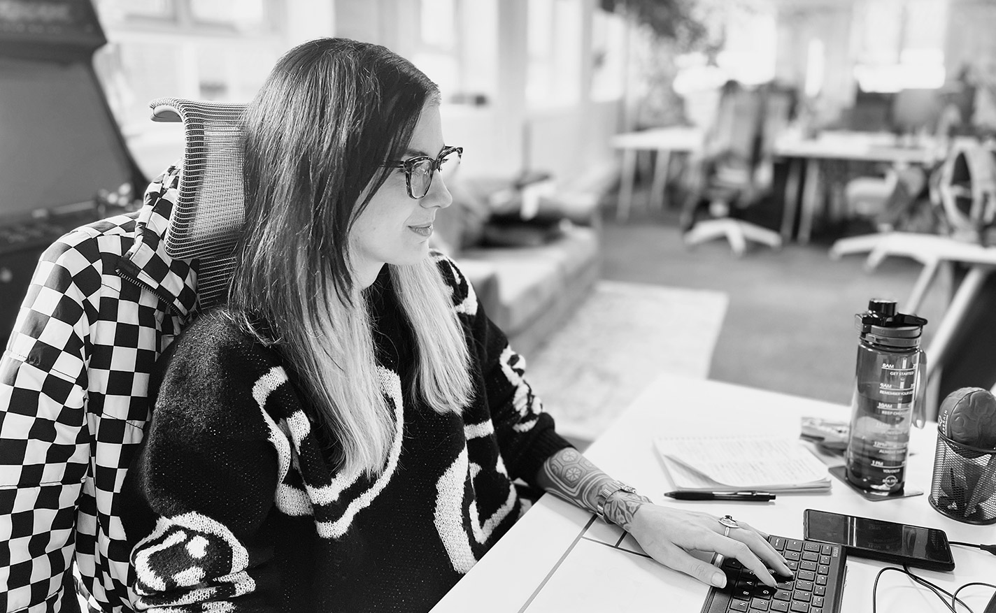 A woman with glasses and tattoos works on a computer at a desk, with a notebook, pen, and water bottle nearby in a modern office setting.