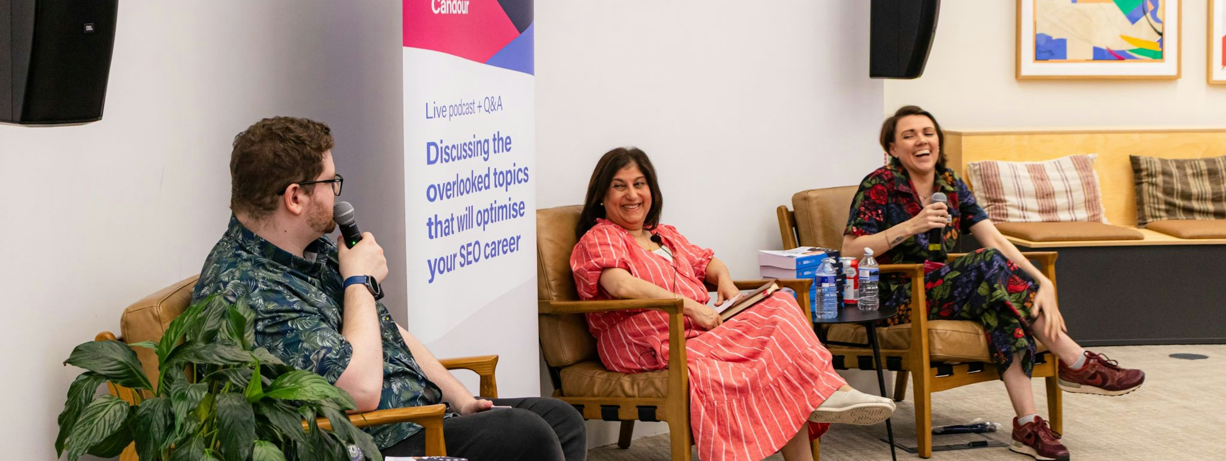 Jack Chambers-Ward, Tazmin Suleman and Sarah McDowell are sitting in a row in comfortable looking armchairs. They are hosting a live podcast and smiling as they talk to each other while holding microphones.